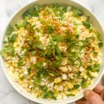 A hand holds a white bowl filled with Mexican street corn salad, topped with crumbled cheese, chopped cilantro, green onions, and sprinkled with chili powder on a white marble surface.