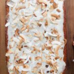 A rectangular coconut lime cake with white icing and toasted coconut flakes on top, placed on a wooden surface.