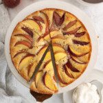 A hand serves a slice of Picnic Cake from a round, golden-brown cake topped with peach slices. Nearby are whole peaches, a bowl of whipped cream, a striped towel, and a pitcher on a light surface.