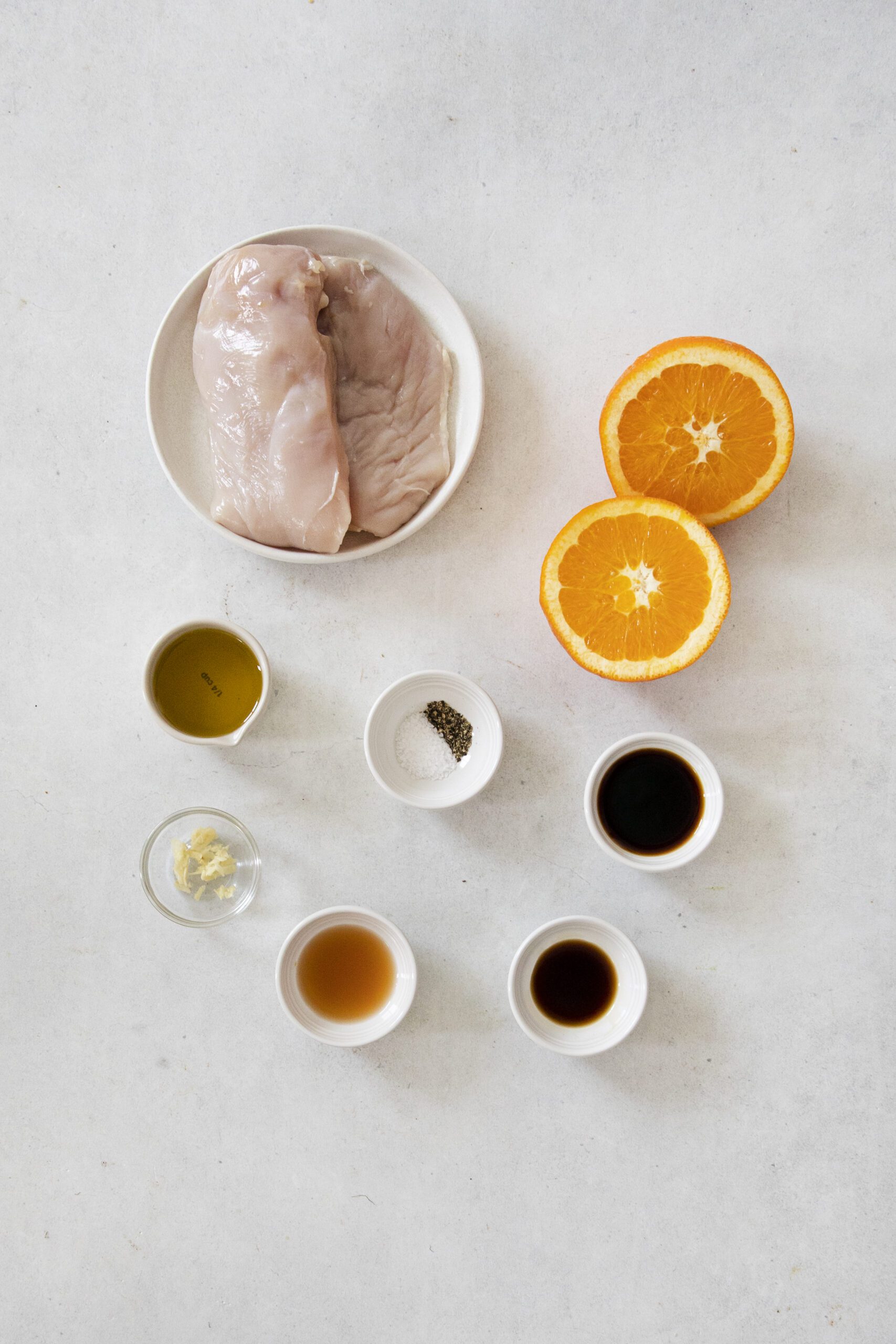 A flat lay of raw chicken breasts on a plate, two orange halves, and small bowls of olive oil, minced garlic, salt and pepper, soy sauce, vinegar, and Worcestershire sauce—perfect ingredients for a flavorful grilled chicken marinade.