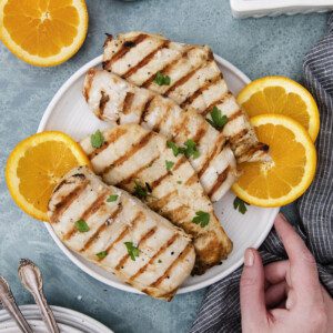 A plate of chicken breasts prepared with a flavorful grilled chicken marinade, garnished with parsley and orange slices, surrounded by fresh orange halves, cherry tomatoes, salt, and a hand holding the plate on a blue surface.