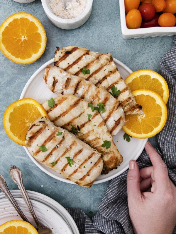 A plate of chicken breasts prepared with a flavorful grilled chicken marinade, garnished with parsley and orange slices, surrounded by fresh orange halves, cherry tomatoes, salt, and a hand holding the plate on a blue surface.