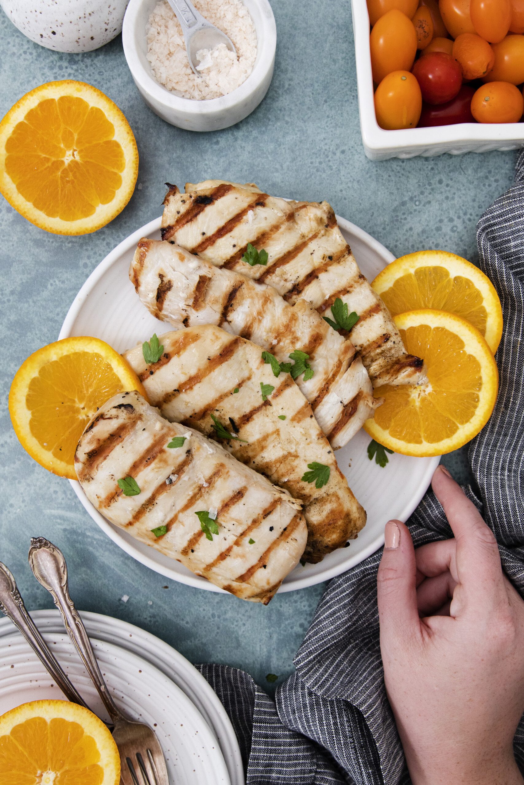 A plate of chicken breasts prepared with a flavorful grilled chicken marinade, garnished with parsley and orange slices, surrounded by fresh orange halves, cherry tomatoes, salt, and a hand holding the plate on a blue surface.