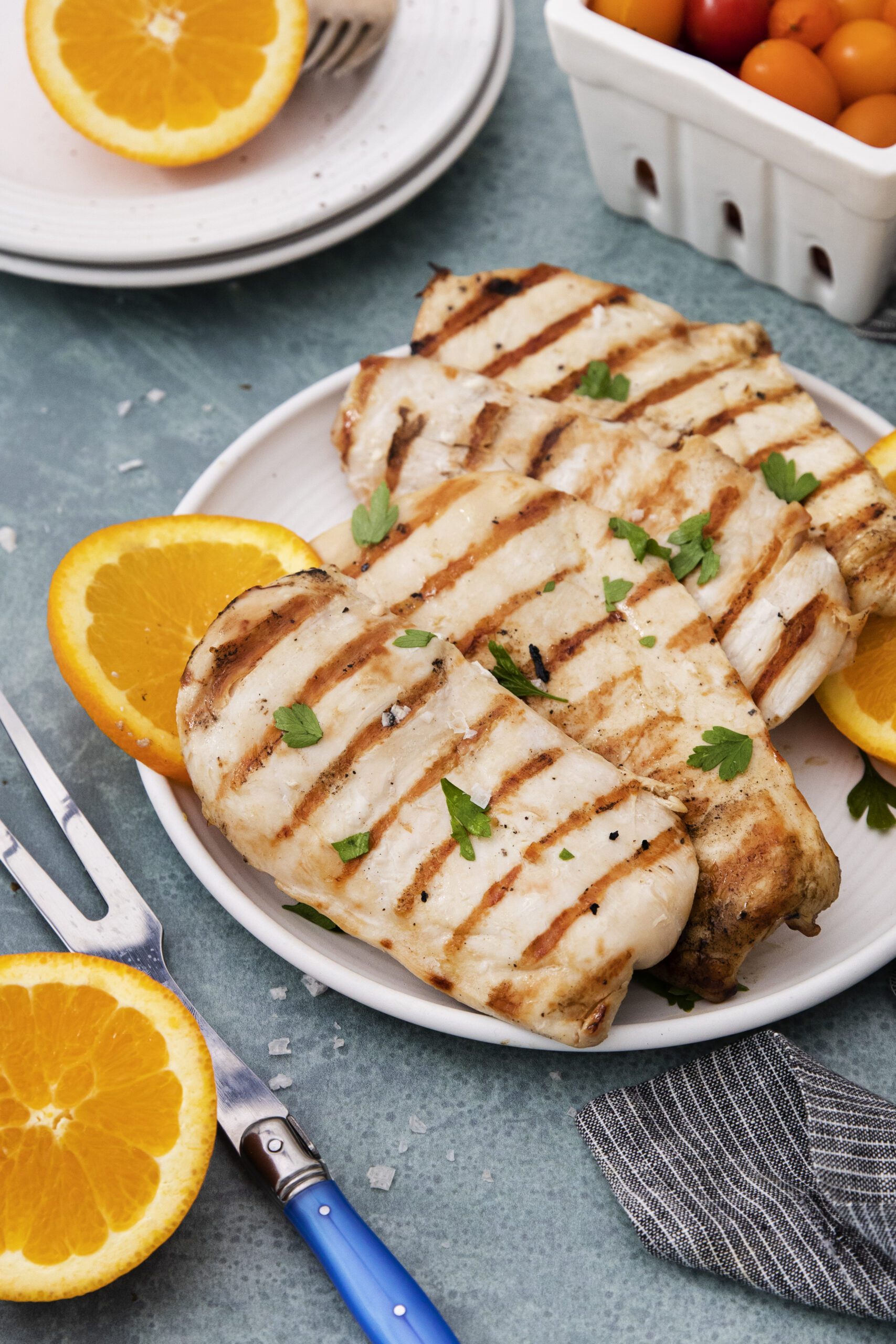 A white plate with grilled chicken breasts, infused with a flavorful grilled chicken marinade and garnished with parsley, is accompanied by orange slices. Cherry tomatoes in a white container and orange halves rest nearby on a blue surface.