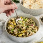A hand holds a fork over a bowl of Mayo-Free Tuna Salad with chopped vegetables. In the background, there is a bowl of round crackers and a small dish of sliced pickled peppers.