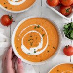 A hand holds a bowl of Creamy Roasted Tomato Soup garnished with a swirl of cream and chopped herbs. Fresh tomatoes and parsley are placed nearby on a white tiled surface.