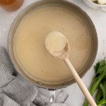 A saucepan filled with creamy white sauce being stirred with a wooden spoon, surrounded by fresh celery, onions, a glass jar of broth, and a bowl of flour—perfect ingredients for make-ahead gravy on a light surface.