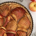 A baked apple tart with fanned apple slices in a crust, sprinkled with brown sugar. The tart is in a round pan on a light surface, beside a cup of brown sugar and two whole apples, highlighting the classic appeal of an apple tart.
