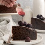 A hand drizzles cherry sauce from a spoon onto a slice of chocolate olive oil cake topped with cherries, placed on a white plate with a fork. A whole cake and white bottles are in the background.