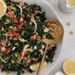 A bowl of Tuscan Kale Salad with chopped tomatoes, fennel, walnuts, and chickpeas, garnished with lemon wedges. A gold spoon rests in the salad. Nearby are lemon halves, a small pitcher of dressing, and a bowl of chickpeas.