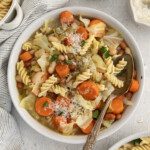 A bowl of Vegetable Minestrone Soup with rotini pasta, carrots, cabbage, and white beans, topped with grated cheese. A vintage spoon rests in the bowl, surrounded by bowls of fresh parsley, grated cheese, and a striped napkin.