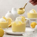 A hand holds a spoonful of yellow Lemon Meringue Pie Pudding above a glass cup topped with whipped cream and lemon slice, surrounded by lemons and more mousse cups on a light table.