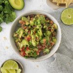 A bowl of chunky guacamole with diced tomatoes and green onions, surrounded by tortilla chips, avocado halves, lime wedges, and fresh cilantro on a light surface.