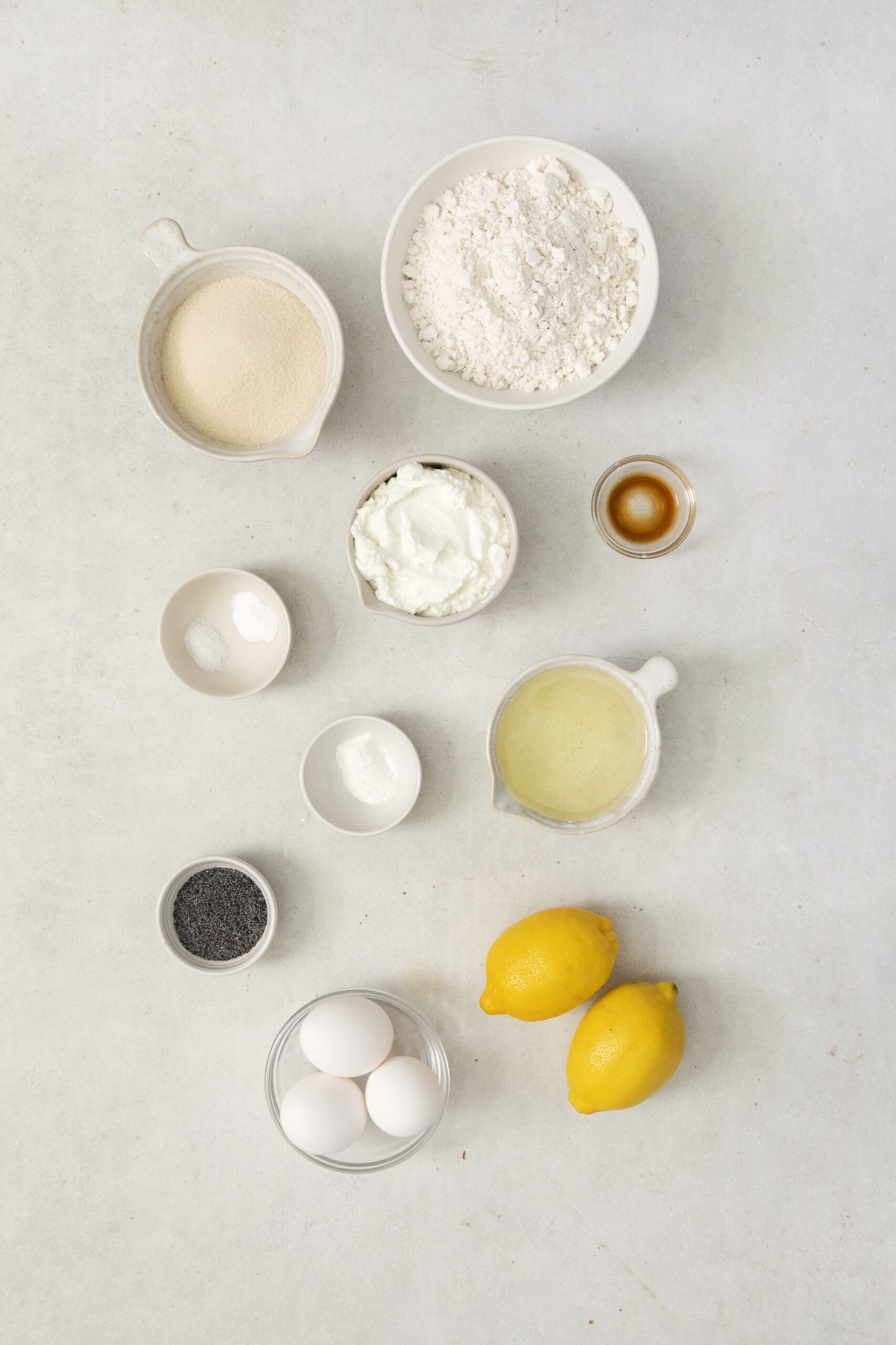 Overhead view of baking ingredients for Lemon Poppy Seed Muffins on a light surface, including flour, sugar, yogurt, eggs, poppy seeds, lemon juice, baking powder, baking soda, vanilla extract, and two whole lemons.