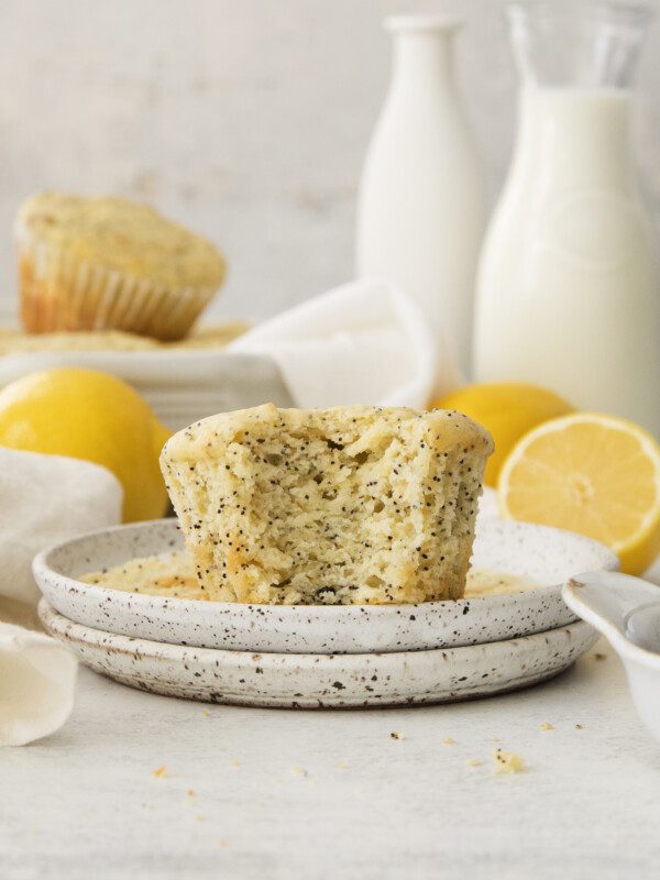 A Lemon Poppy Seed Muffin with a bite taken out sits on a speckled plate, surrounded by halved and whole lemons, milk jars, and another Lemon Poppy Seed Muffin in the background.