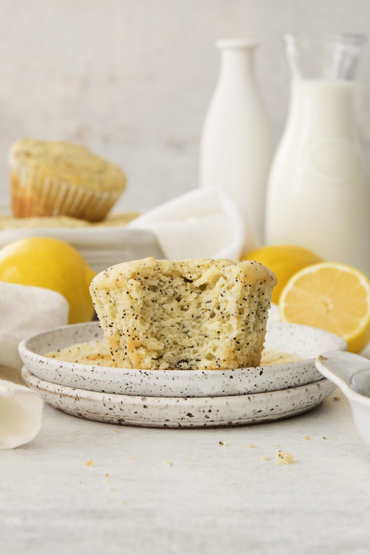 A Lemon Poppy Seed Muffin with a bite taken out sits on a speckled plate. Whole lemons, a lemon half, another muffin, and bottles of milk surround the scene on a light surface.