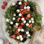 A platter of Berry Caprese Salad with arugula, fresh strawberries, blackberries, mozzarella balls, and sliced shallots. Two gold serving utensils and a pair of hands hold the plate. A strawberry, mint, and plates are nearby.