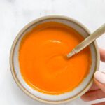 A hand holds a bowl filled with bright orange hot sauce, with a spoon resting inside, on a white marble surface.