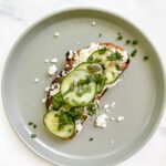 Ricotta Toast topped with creamy ricotta cheese, crisp cucumber slices, chopped green onions, and fresh herbs sits on a light gray plate. Crumbled cheese and green onions are artfully scattered around for a fresh finish.