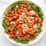 A vibrant Arugula and Blistered Tomato Salad featuring roasted cherry tomatoes, white beans, sliced shallots, and fresh herbs, beautifully arranged on a white marble surface.