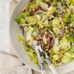 A bowl of Famous La Scala Chopped Salad with lettuce, chickpeas, shredded cheese, and sliced salami sits ready with serving utensils. A small bowl of chickpeas rests nearby on a beige cloth napkin.