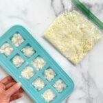 A hand holds a blue ice cube tray filled with frozen cauliflower rice cubes—perfect for batch prep like when you mince and freeze garlic—next to a resealable plastic bag containing loose cauliflower rice on a white marble surface.