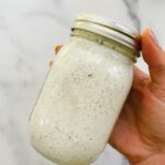 A hand holds a mason jar filled with creamy Miso-Ranch Dressing, speckled with green herbs, against a white marble background.