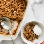 A hand holds a bowl of Cardamom Apple Crisp topped with vanilla ice cream, next to a baking dish filled with more Cardamom Apple Crisp. A serving spoon rests in the dish on a white marble surface.