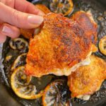 A hand holds a crispy, golden-brown chicken thigh above a skillet filled with more crispy chicken thighs and browned lemon slices.