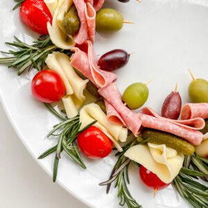 A Holiday Charcuterie Wreath on a white plate features rolled salami, cheese slices, cherry tomatoes, green and black olives, mini pickles, and rosemary sprigs for a festive and delicious centerpiece.