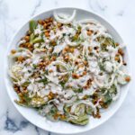 A white plate on a marble surface holds a Caramelized Cabbage Salad with sliced fennel, crispy chickpeas, fresh herbs, and a creamy white dressing drizzled on top.