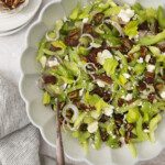 A scalloped bowl filled with Celery Chopped Salad, topped with pecans, sliced shallots, feta cheese, and dates. Nearby are small dishes of pecans and feta, and a striped cloth is draped on the table.