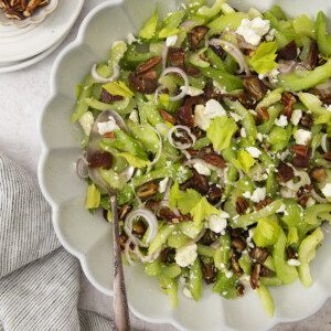 A scalloped bowl filled with Celery Chopped Salad, topped with pecans, sliced shallots, feta cheese, and dates. Nearby are small dishes of pecans and feta, and a striped cloth is draped on the table.