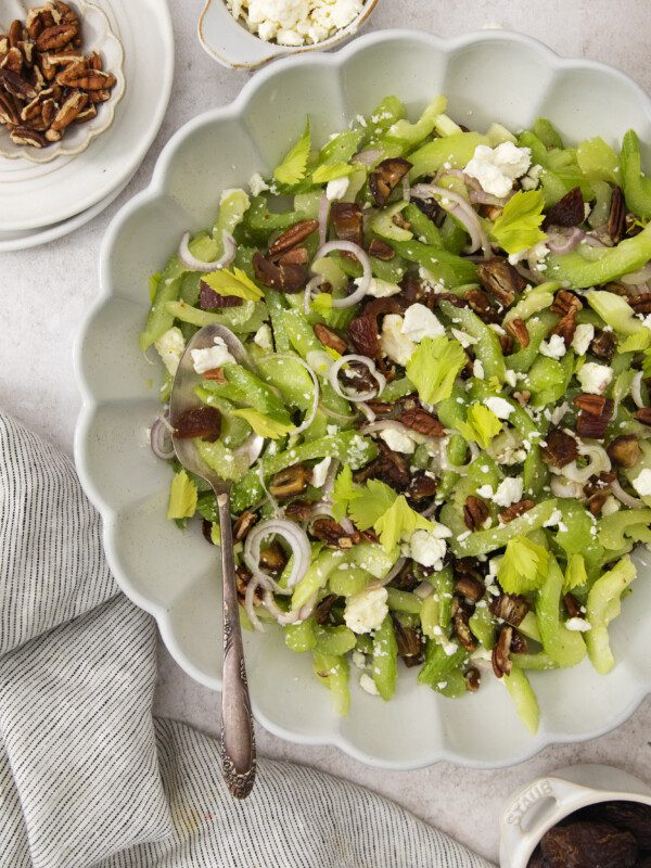 A scalloped bowl filled with Celery Chopped Salad, topped with pecans, sliced shallots, feta cheese, and dates. Nearby are small dishes of pecans and feta, and a striped cloth is draped on the table.