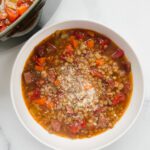 A bowl of Lentil Sausage Soup with Fried Garlic, featuring diced carrots, celery, tomatoes, and sausage, topped with grated cheese, sits on a white surface next to a pot of the same soup.