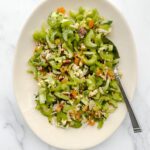 A white oval plate with a fresh Celery Chopped Salad, topped with chopped nuts, dried apricots, and herbs, with a fork resting on the side. The background is a white marble surface.
