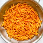 A stainless steel pan filled with Creamy Roasted Red Pepper Pasta—penne coated in a luscious orange sauce—viewed from above on a white surface.