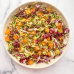 A bowl of colorful Shaved Brussels Sprouts and Butternut Squash Salad with chopped greens, purple radicchio, and orange butternut squash sits on a white marble surface.