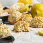 Lemon Coconut Macaroons on a light surface, with a bite taken out of one. Lemons, lemon slices, and a glass of milk sit in the background, creating a scene that looks bright and fresh.