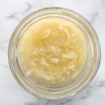 A glass bowl filled with tangy shredded sauerkraut and its brine, placed on a white marble surface.