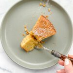 A hand holds a fork with a bite of Maialinos Olive Oil Cake, its crumbly, golden texture dusted with powdered sugar on a light gray plate set on a marble surface.