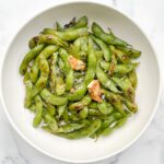 A white bowl filled with blistered edamame pods, topped with coarse salt, rosemary, and roasted garlic, set on a marble surface.