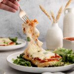 a hand holding a fork, pulling a string of cheese from baked chicken parm