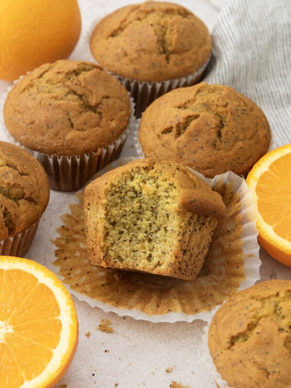 A group of Orange Poppy Seed Muffins sits on a light surface with whole and halved oranges nearby. One muffin, with a bite taken out, is unwrapped to reveal its soft, speckled interior. A striped cloth is in the corner.