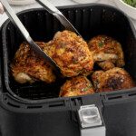 A hand uses metal tongs to lift a piece of seasoned, cooked Air Fryer Chicken Thighs from a basket filled with golden-brown pieces. Plates and fresh herbs are visible in the background.
