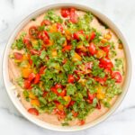 A bowl of 7-Layer Dip topped with chopped tomatoes, yellow bell peppers, cilantro, and green onions sits on a white marble surface.