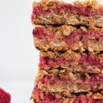 A close-up of four stacked Mixed Berry Crumble Bars showcases layers of oats and vibrant raspberry filling; a single raspberry sits to the side on a plain white background.