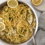 A pan of Artichoke Scampi Pasta, garnished with chopped herbs and lemon slices, sits on a light surface. A spoon rests in the pan, with halved lemons, stacked plates, and a gray checked napkin nearby.