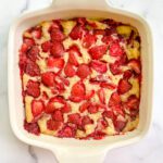 A Strawberry Dump-It Cobbler baked in a square white dish, with a golden crust and sliced strawberries on top, sits invitingly on a white marble surface.