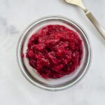 A glass bowl filled with chunky red cranberry sauce sits on a white surface, reminiscent of a vibrant rhubarb chutney, with a metal spoon placed nearby.